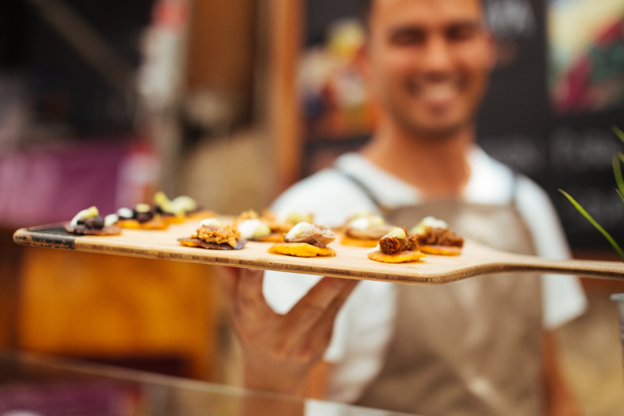 Home Close-up of a smiling chef presenting a tray of gourmet finger foods in a vibrant market setting.